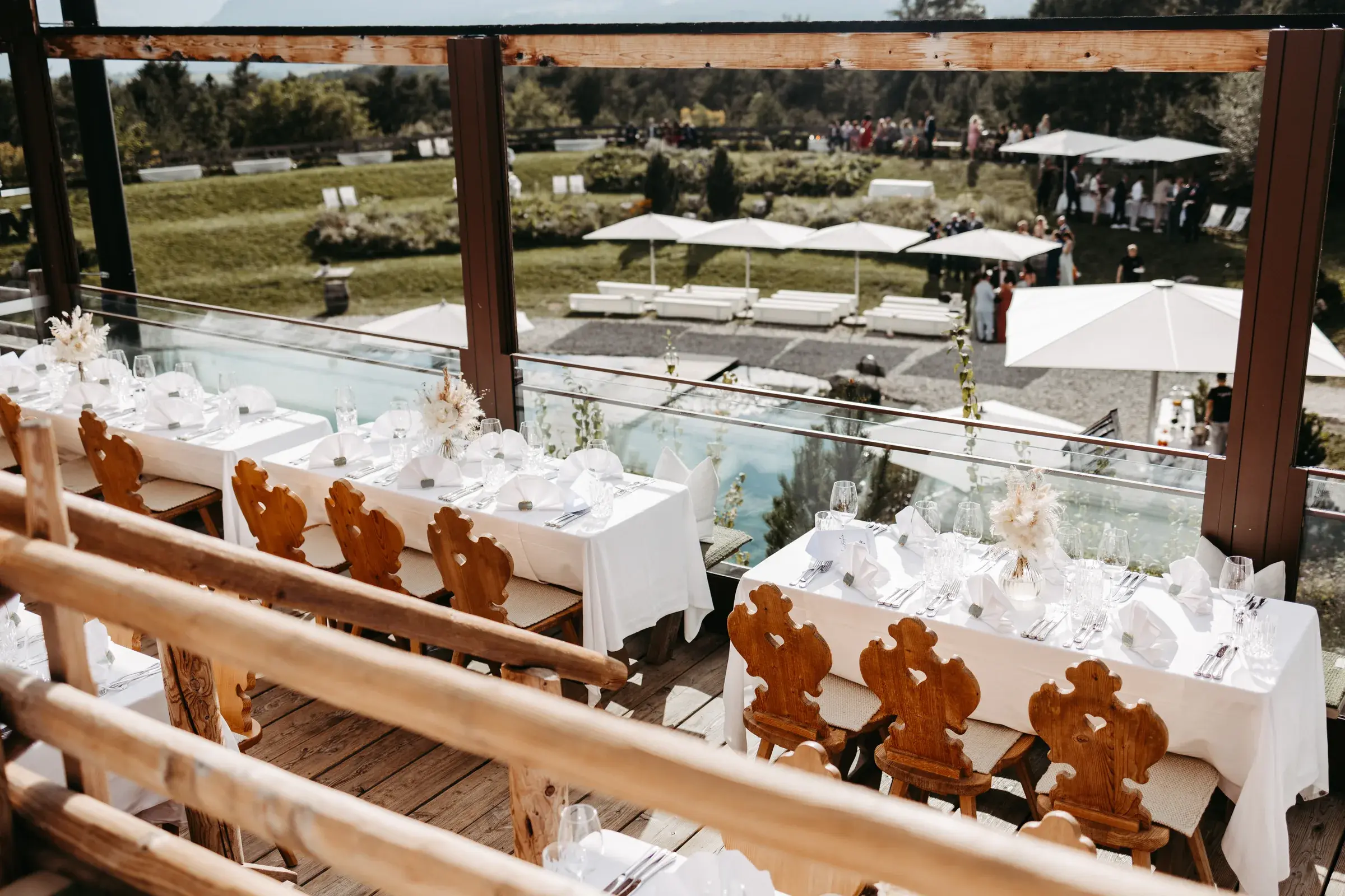 Festively set wedding tables with Tyrolean wooden chairs on the roofed Stöttlalm terrace overlooking pond and mountains