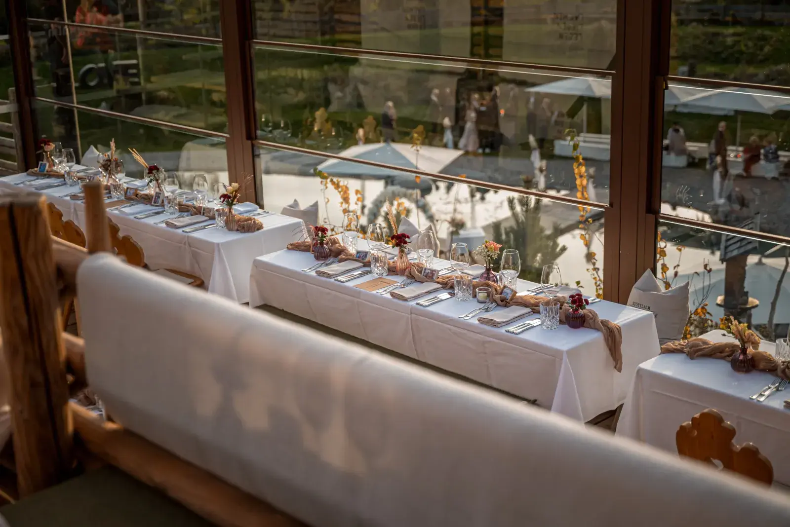 Lange festlich weiß gedeckte Hochzeitstafel mit Trockenblumen und Kristallgläsern entlang der Fensterfront der Stöttlalm, dahinter die Terrasse im Abendlicht